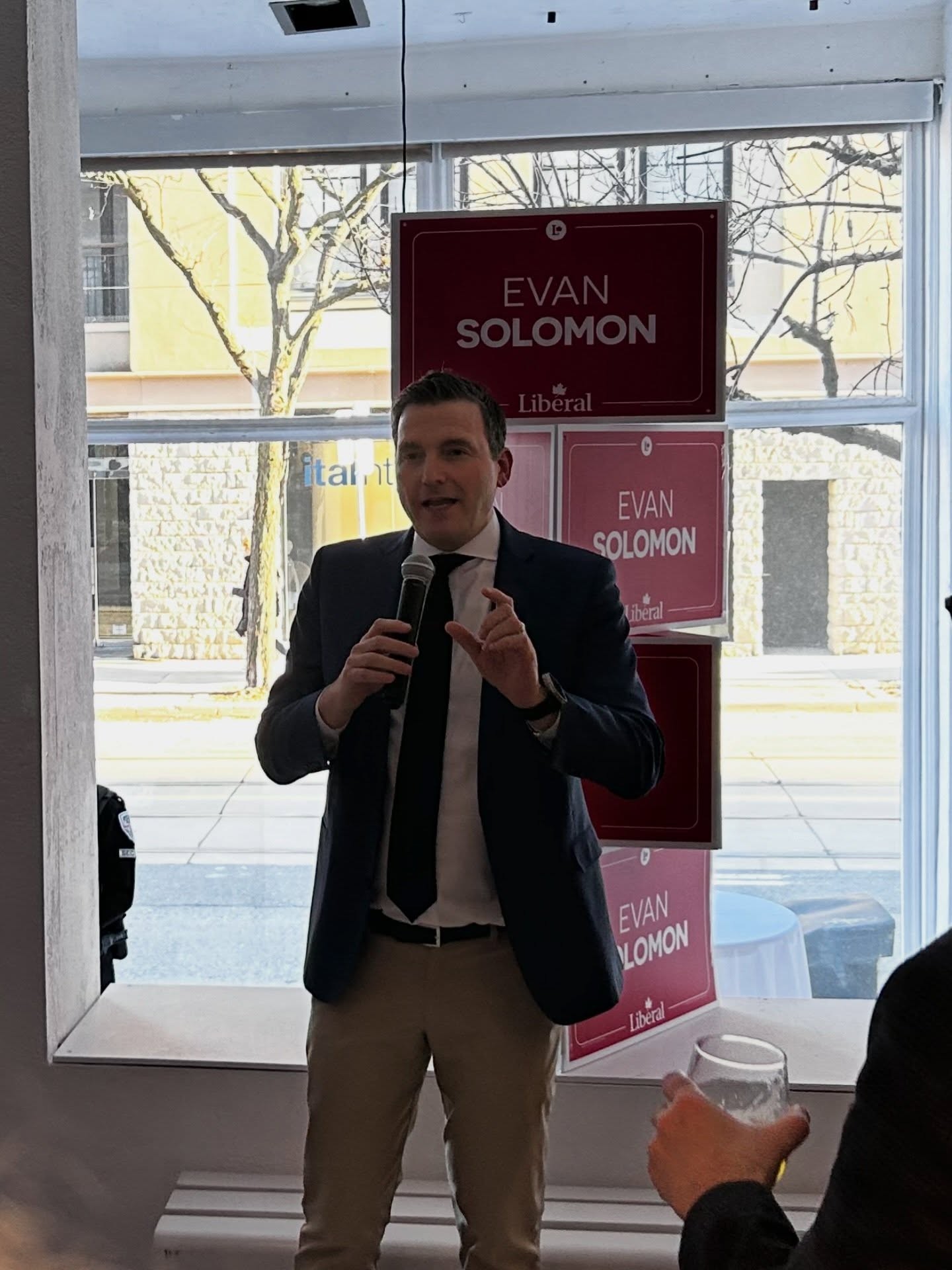 10 - The Canada Times Man in a suit speaks into a handheld microphone at a campaign event, with Evan Solomon Liberal posters in the background.