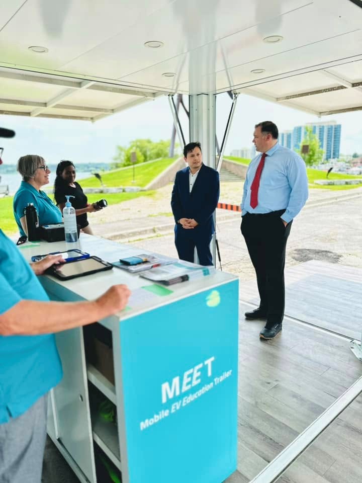 10 - The Canada Times A group chats at a teal information booth labeled MEET Mobile EV Education Trailer under a canopy outdoors.