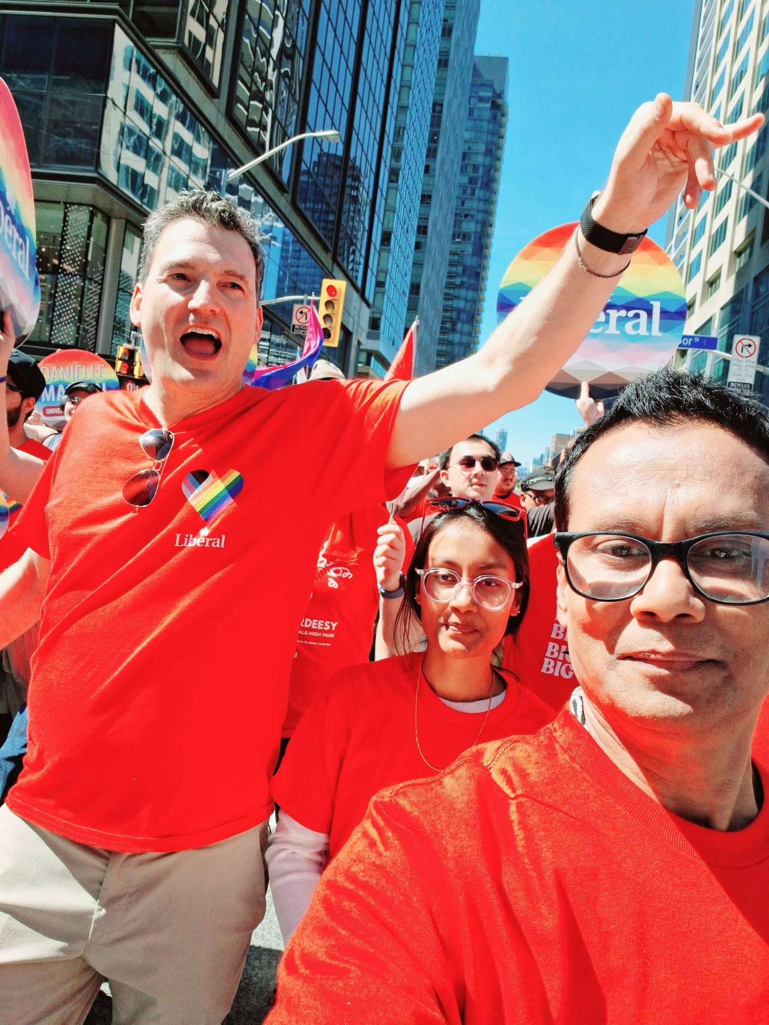 Group of people wearing red shirts at a Pride parade, smiling and posing for a selfie in a city street with rainbow decorations