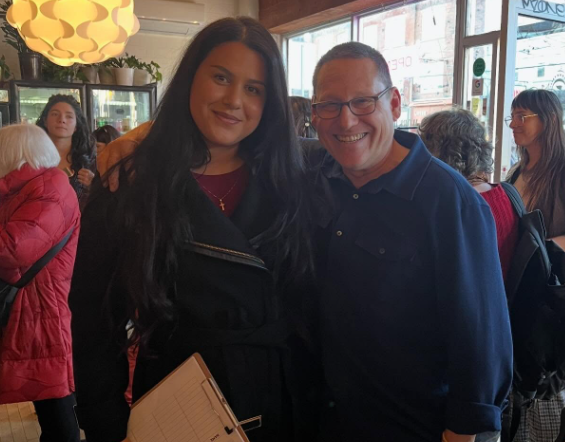 11 - The Canada Times Smiling young woman in a black coat posing with an older man in a blue polo shirt inside a busy cafe?
