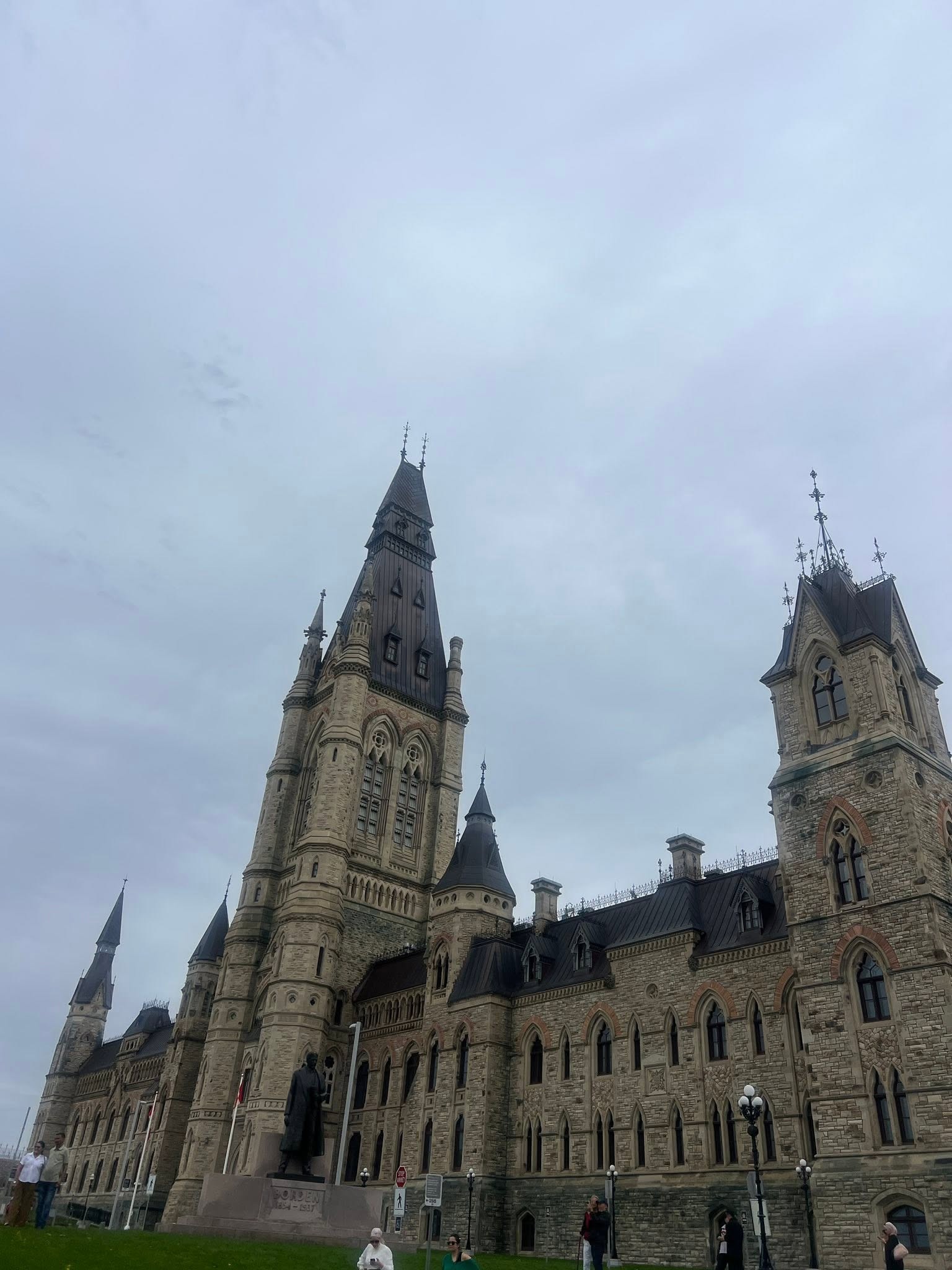 Front facade of a Gothic-style stone government building with tall spires and arched windows, viewed from the lawn with a few visitors nearby.