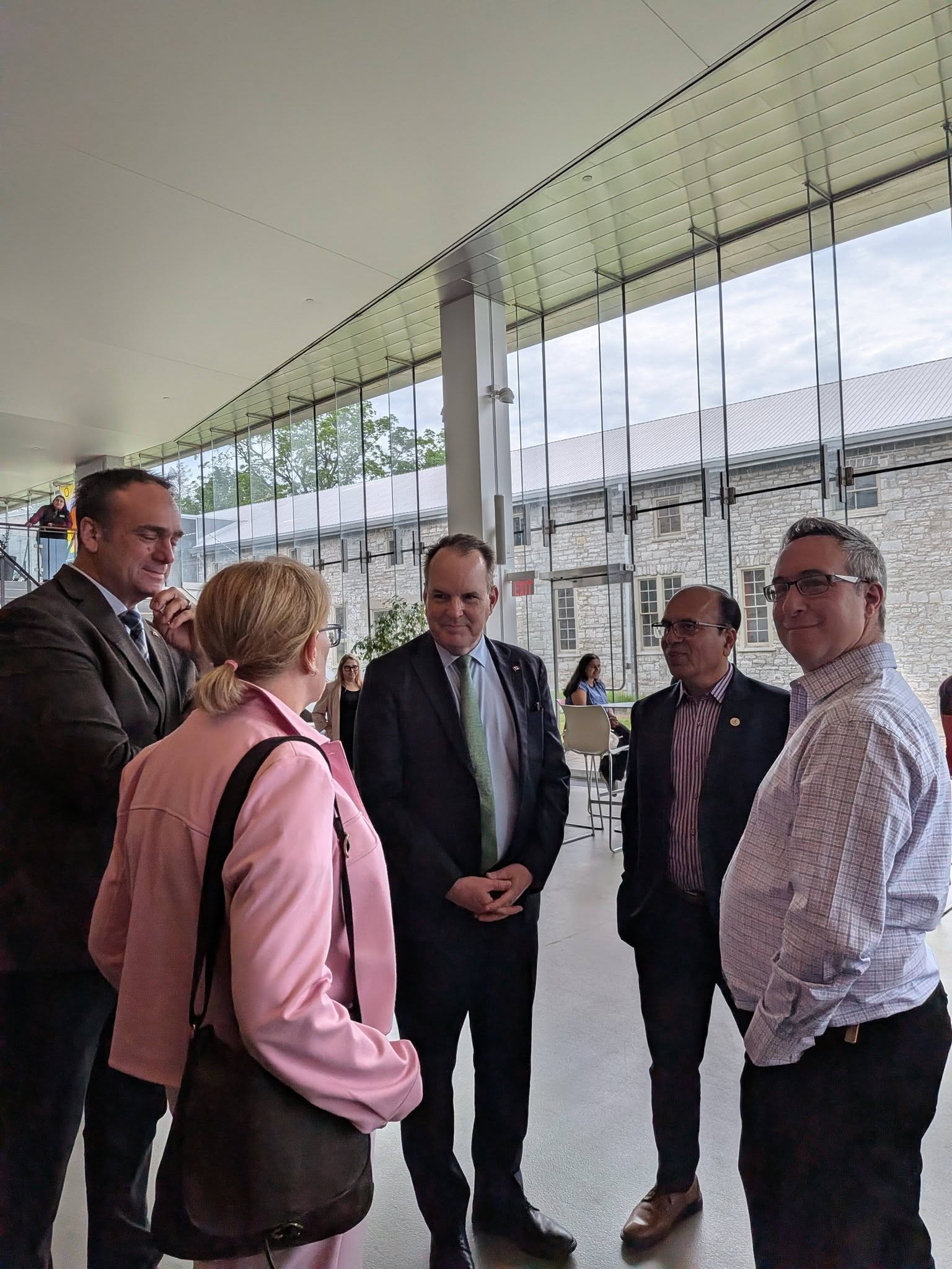 Group of professionals in business attire standing in a glass-walled lobby, chatting.