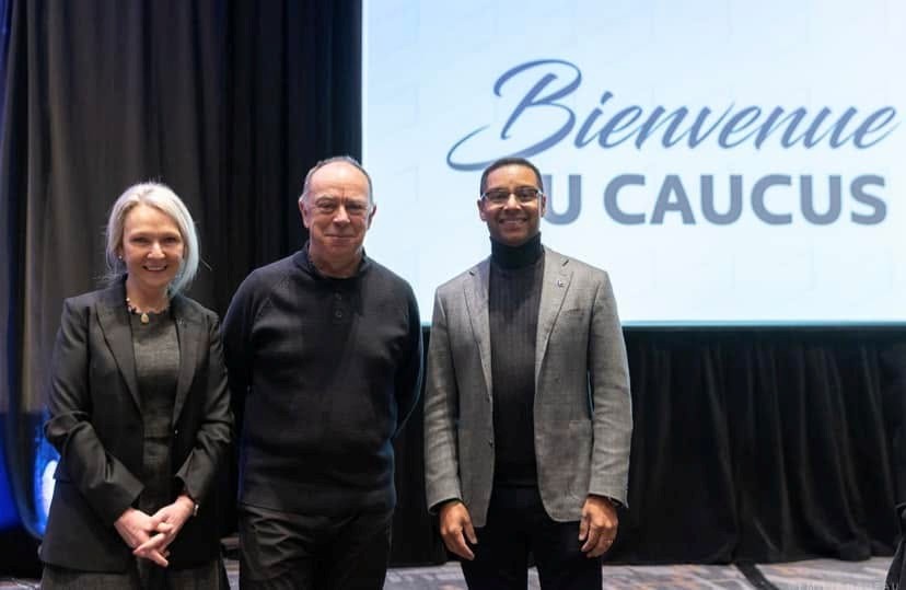 Three people pose on a stage in front of a screen that reads 'Bienvenue au Caucus'.