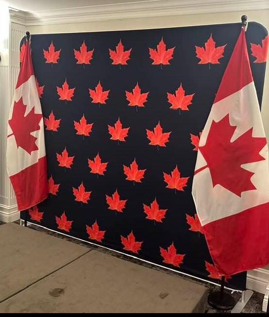 12 - The Canada Times Navy backdrop with repeated red maple leaves, flanked by two Canadian flags indoors.