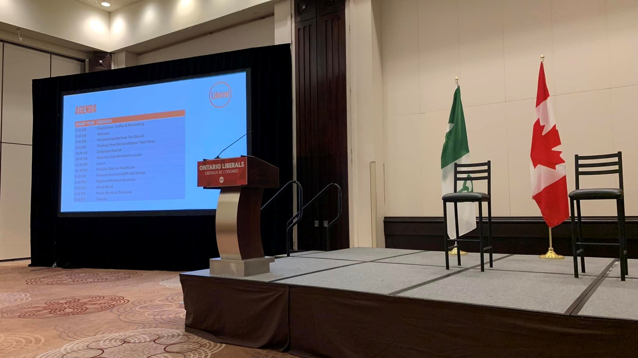 Stage setup for a political event: podium, large screen displaying an agenda and the Ontario Liberals logo, with Canadian and green-and-white flags on stage.