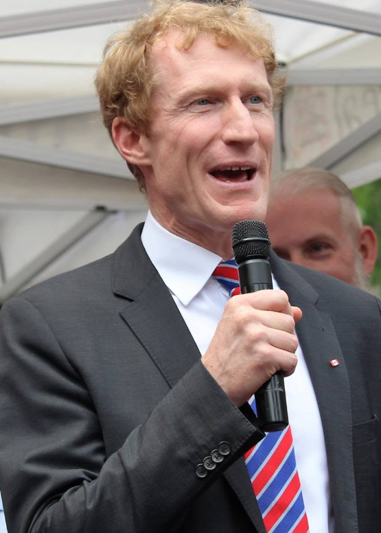 13 - The Canada Times Man in a dark suit and striped red, white, and blue tie speaks into a handheld microphone at an outdoor event, with a tent-like structure in the background.