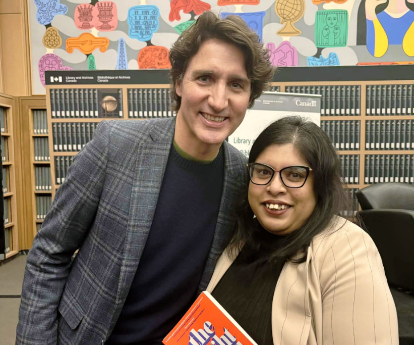 13 - The Canada Times Two people pose for a photo in a library; a man in a blue plaid blazer stands beside a woman with glasses who is holding an orange book.