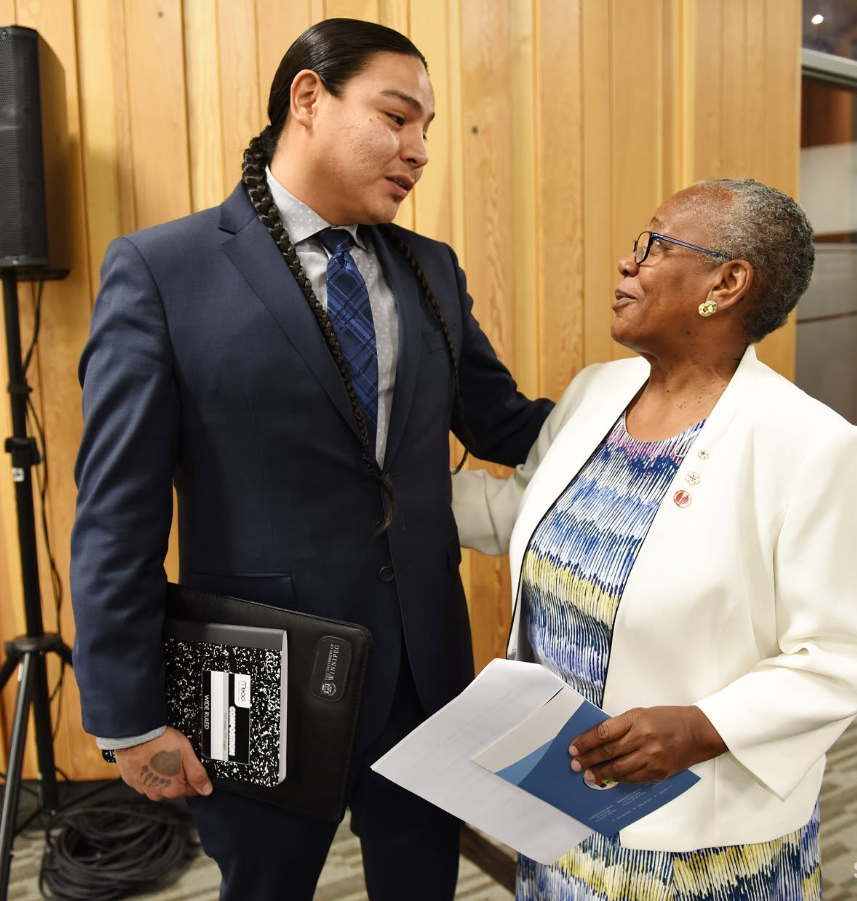 2 - The Canada Times Man in a navy suit with long braided hair talks with an older woman in a white blazer, both holding documents in a wood-panel room behind them.