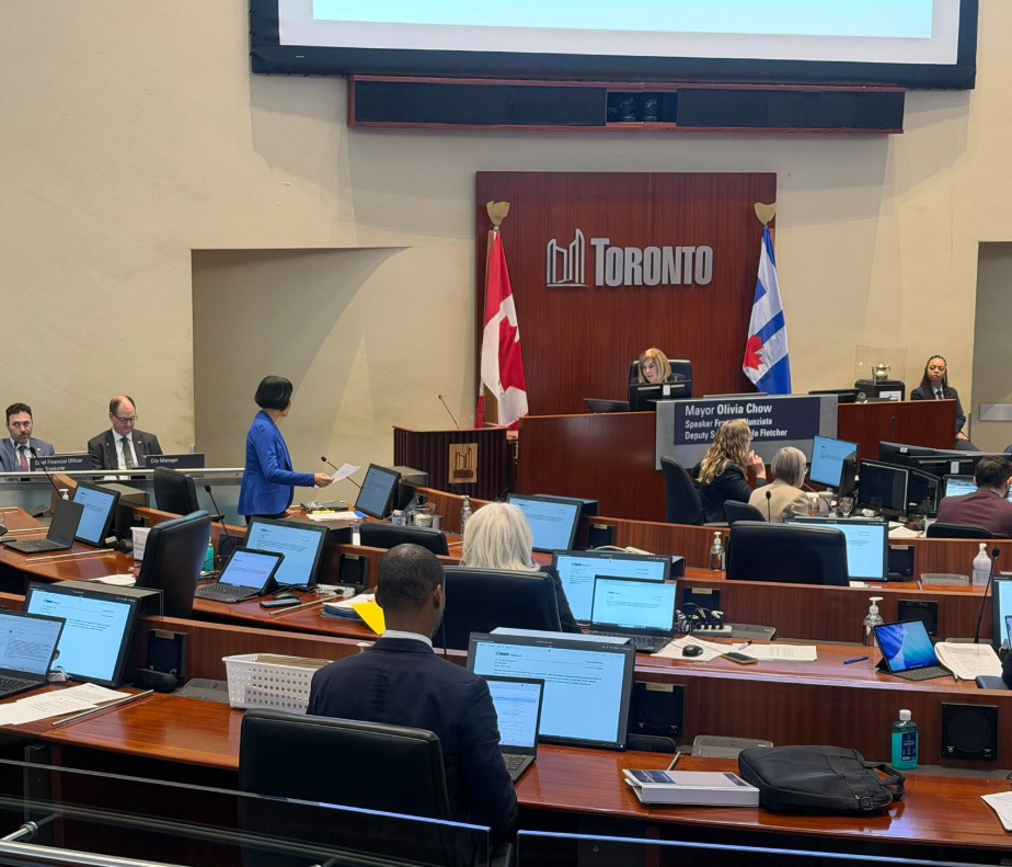 Toronto city council chamber with officials at desks and a speaker at the front podium.