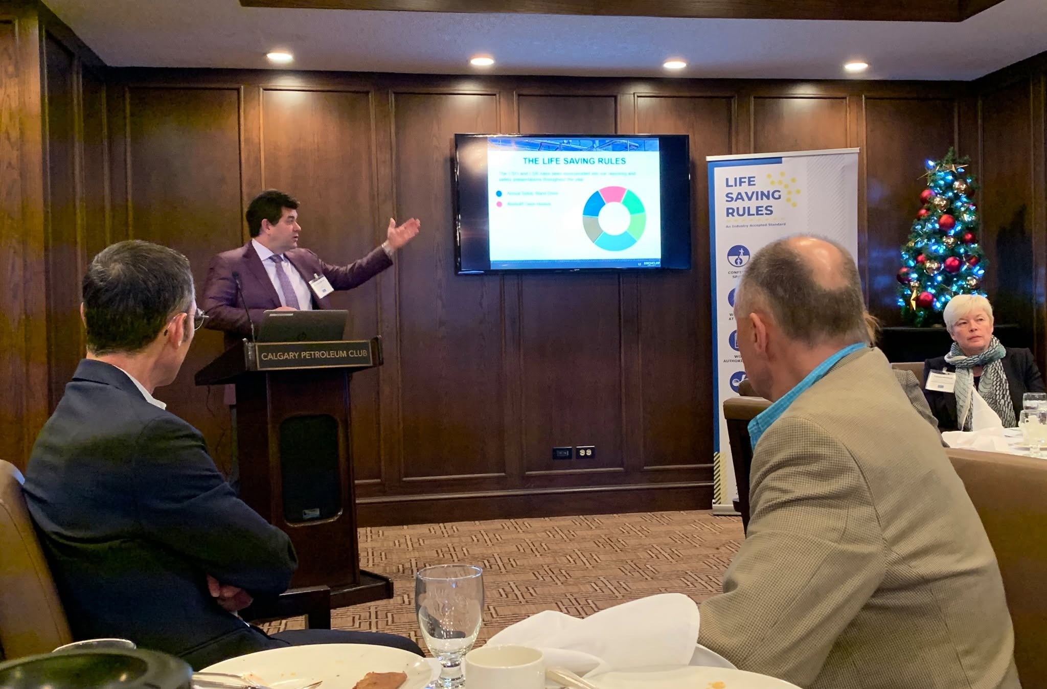 2 - The Canada Times Presenter in a purple suit speaks at a wooden podium in a formal dining room; screen shows 'Life Saving Rules' with a donut chart and bullet points, as attendees listen.