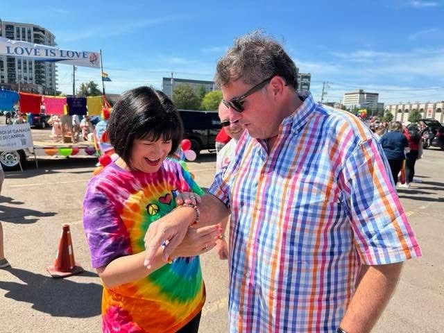 3 - The Canada Times Man helps a smiling woman adjust her bracelet at an outdoor festival; a 'LOVE IS LOVE' banner and colorful crowds in the background, woman in a tie-dye shirt.