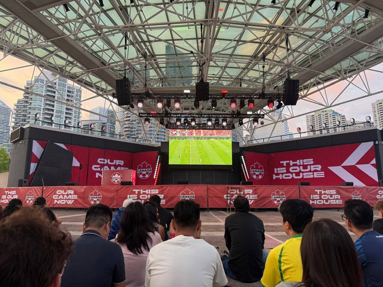 Outdoor stadium viewing area with a large central screen showing a soccer match, surrounded by red banners reading 'THIS IS OUR HOUSE' and 'OUR GAME NOW' and an audience.