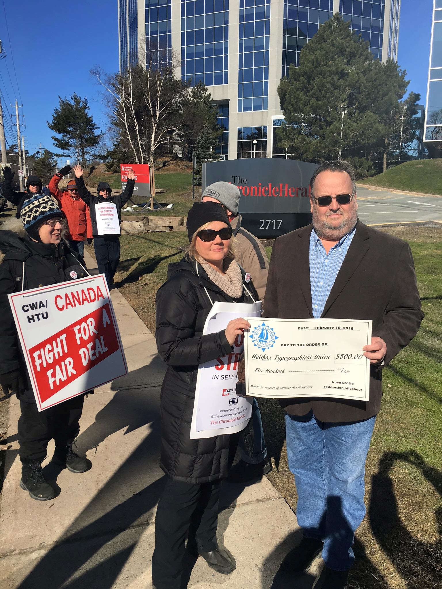 3 - The Canada Times Group of protesters holding signs outside a modern office building; one sign reads 'FIGHT FOR A FAIR DEAL' with 'Canada' visible on others.