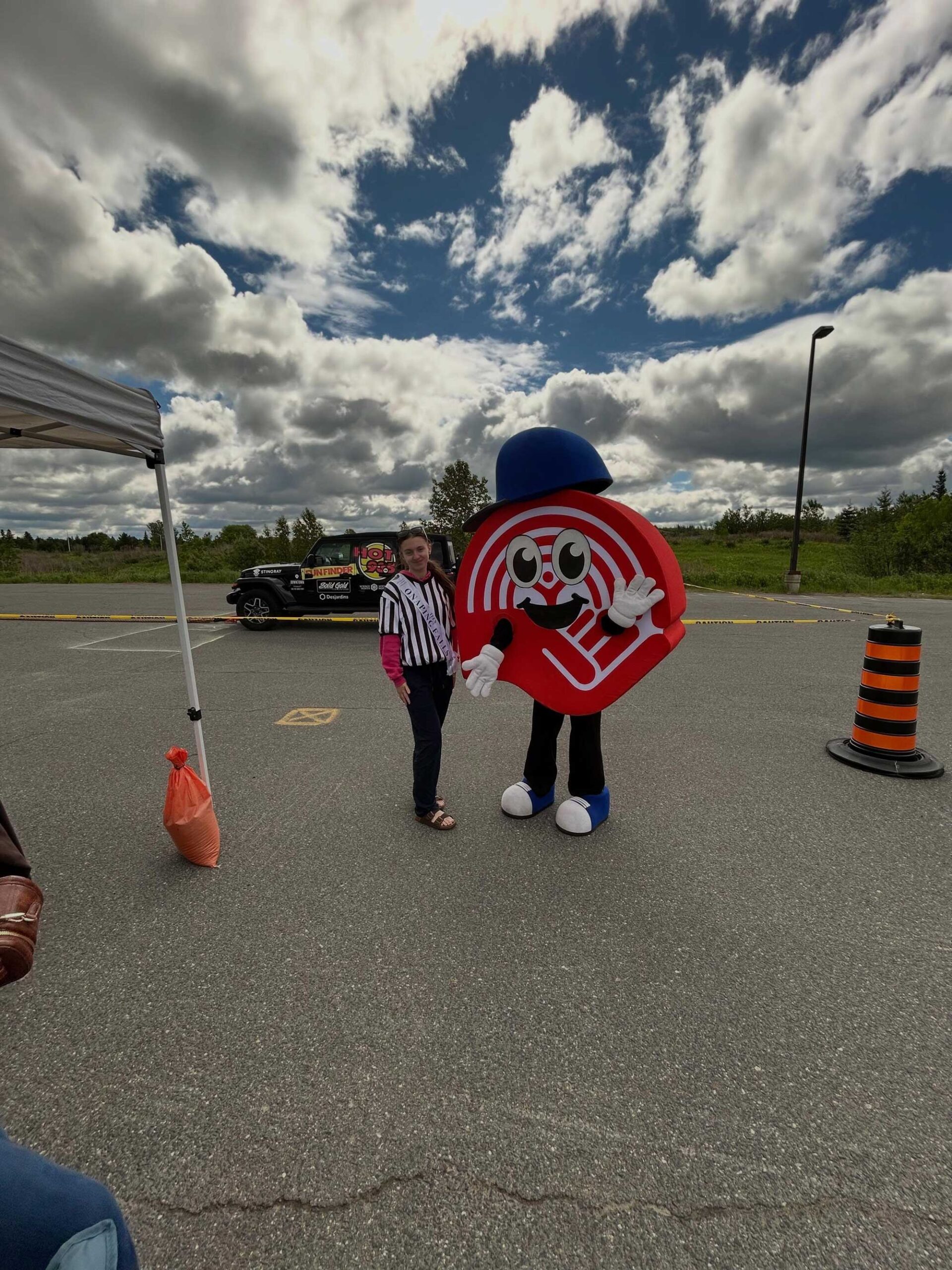Person posing with a large red circular mascot wearing a blue hat in a parking lot under a cloudy sky, with a tent nearby.