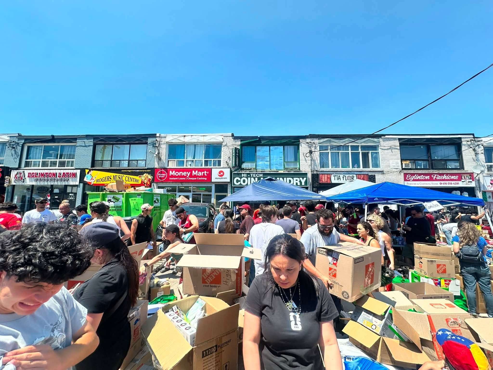 Crowded street market with people rummaging through cardboard boxes in front of storefronts under a clear blue sky.