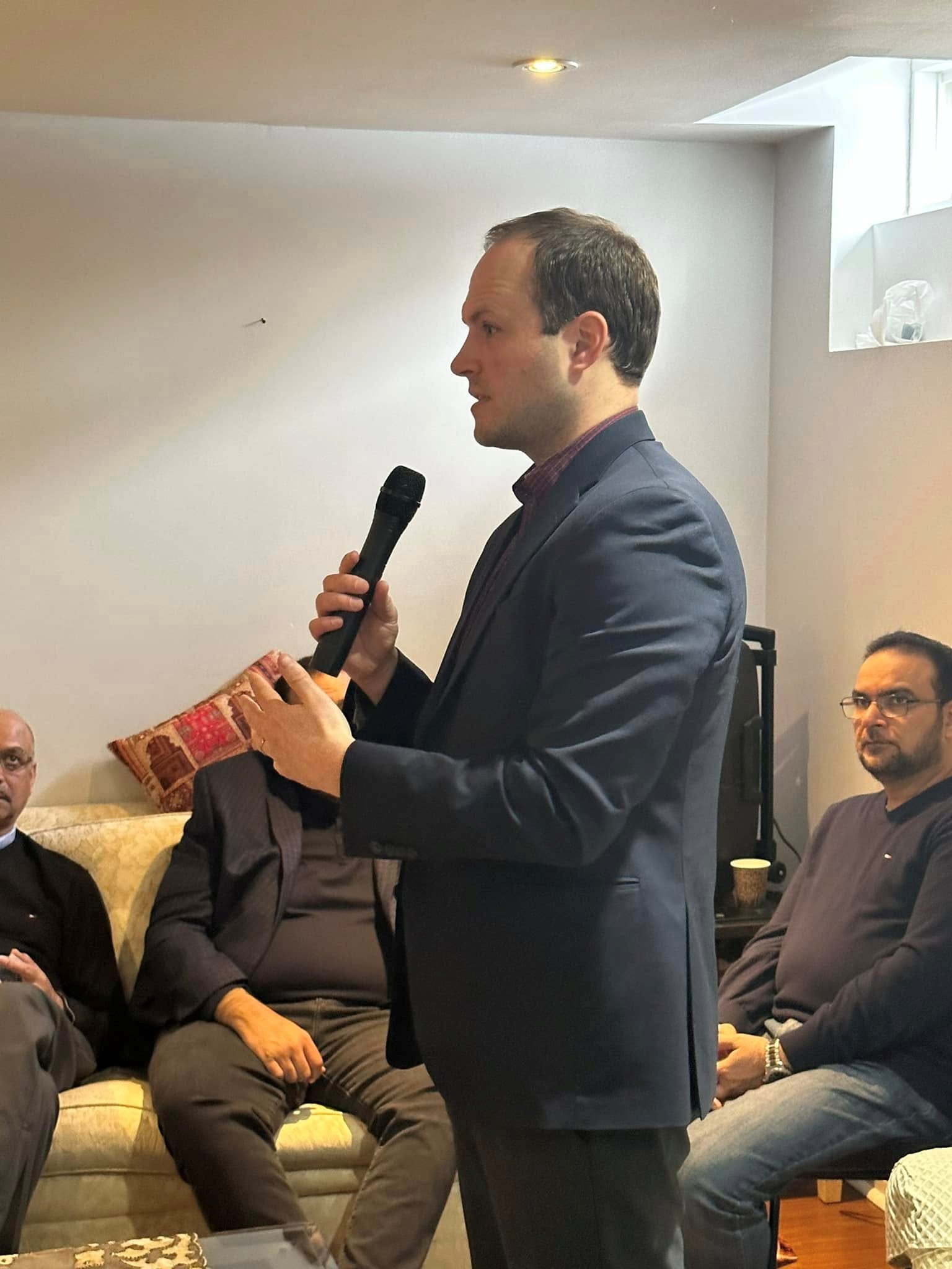 6 - The Canada Times Man in a navy blazer speaking into a handheld microphone at an indoor event, with seated audience in the background.