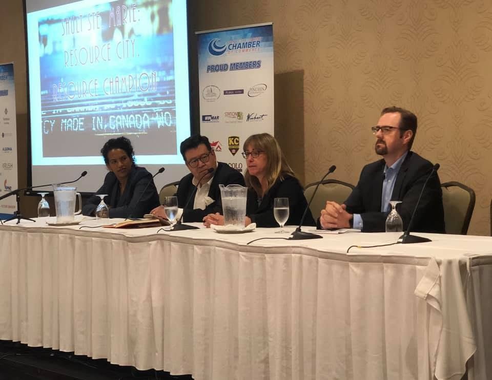 6 - The Canada Times Panel of four speakers at a conference table with microphones; projector screen and banners in the background.