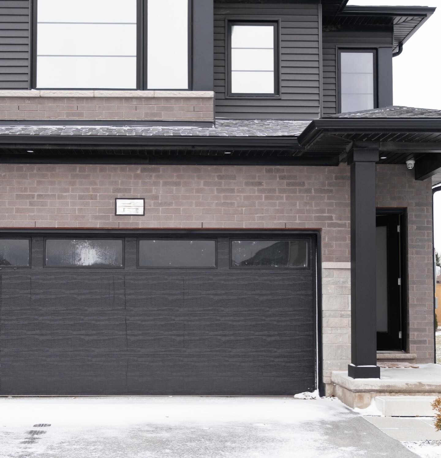 6 - The Canada Times Modern two-story house with dark brick and black siding, a large dark garage door, and a covered entry supported by a black column.