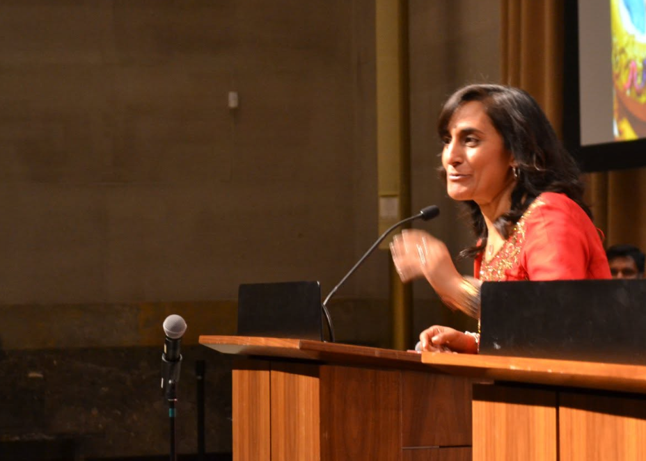 Woman in red traditional outfit speaks at a wooden podium with a microphone, giving a talk.