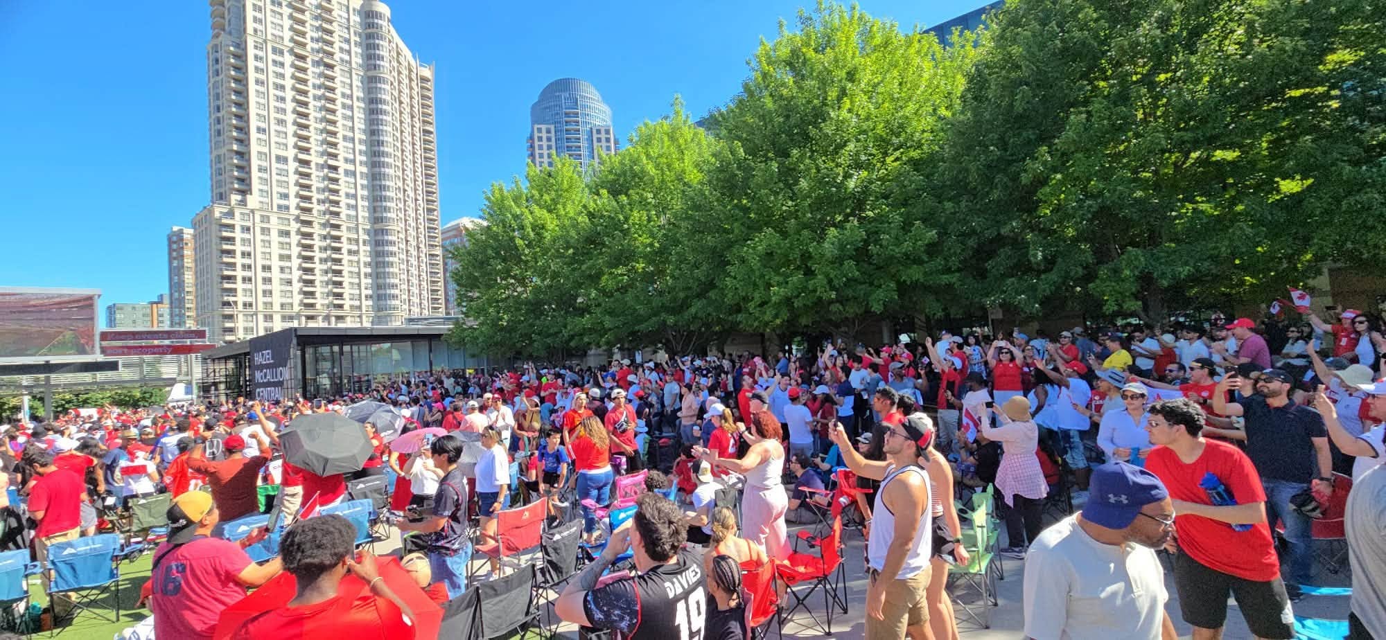Large crowd of people in red shirts gathered in an outdoor plaza under a bright blue sky, with tall modern buildings and green trees behind them.