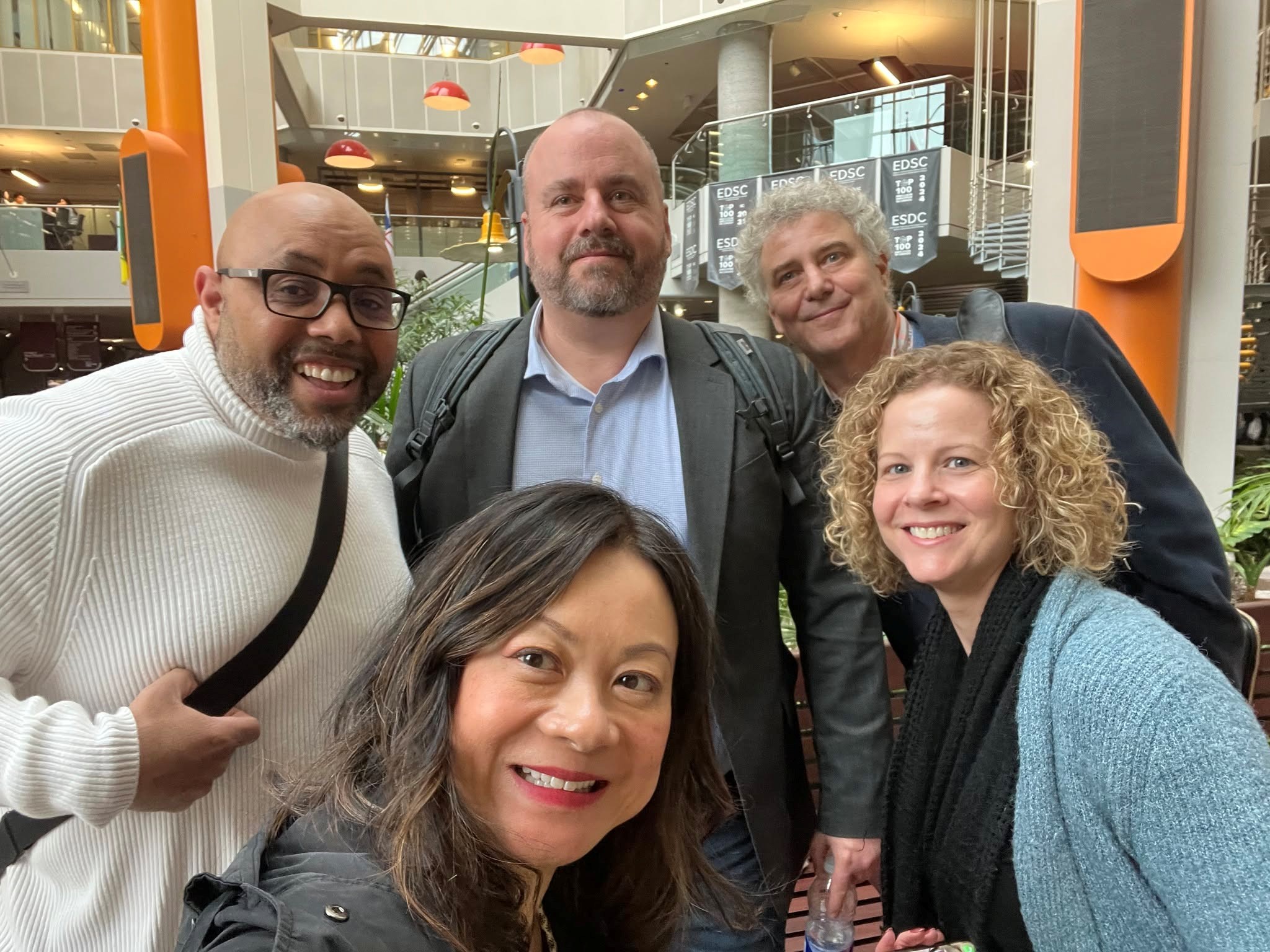 7 - The Canada Times Group of five people smiling for a selfie in a bright, modern atrium with orange pillars and glass railings.