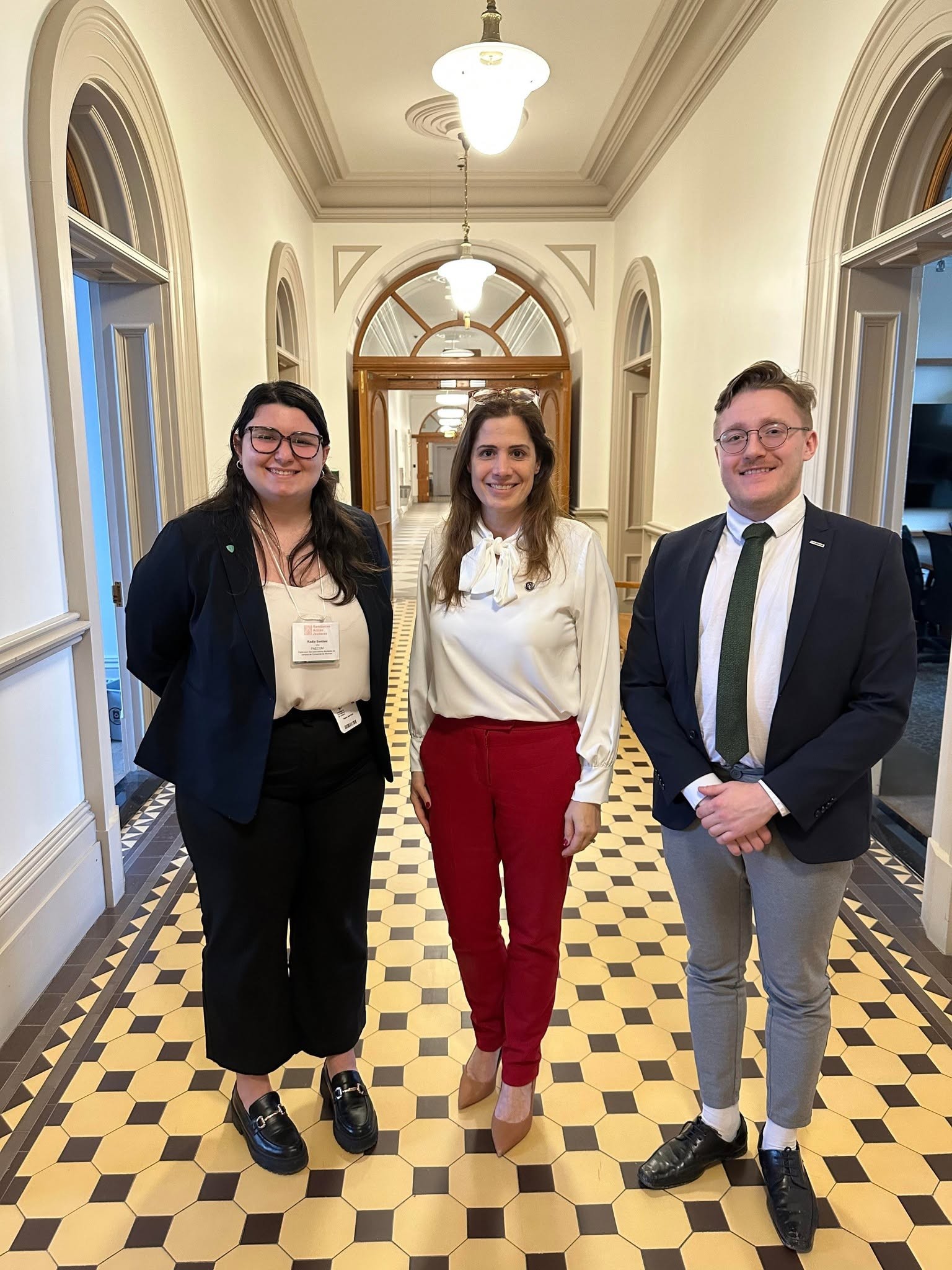 8 - The Canada Times Three professionally dressed people smiling in a bright, arched hallway with checkered floor tiles and wooden doors behind them.