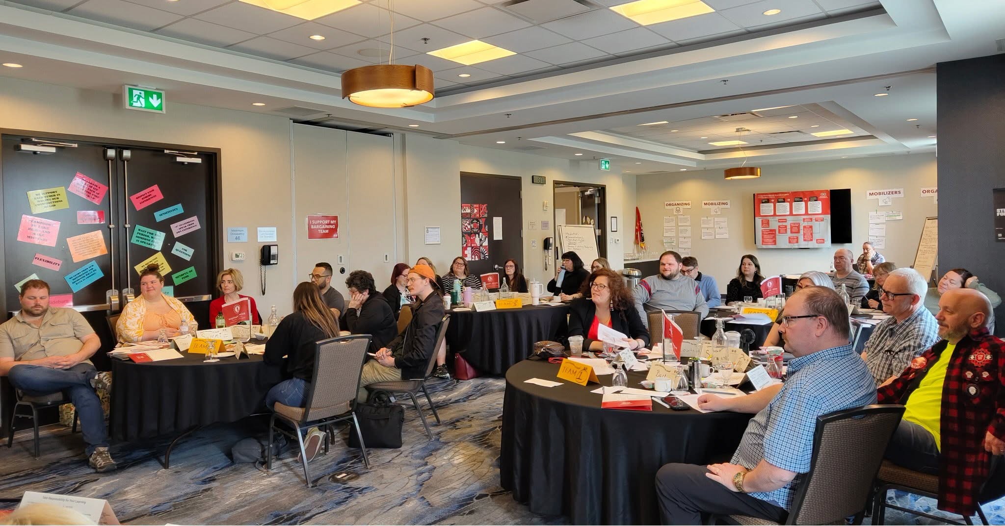 8 - The Canada Times Group of adults seated around round tables in a bright conference room, listening to a presentation with name cards and papers on the tables.