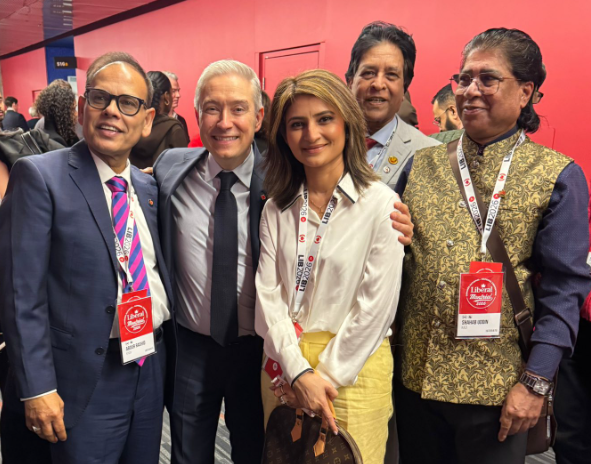 8 - The Canada Times Five people in formal attire pose together at a conference, wearing name badges and lanyards against a pink backdrop.