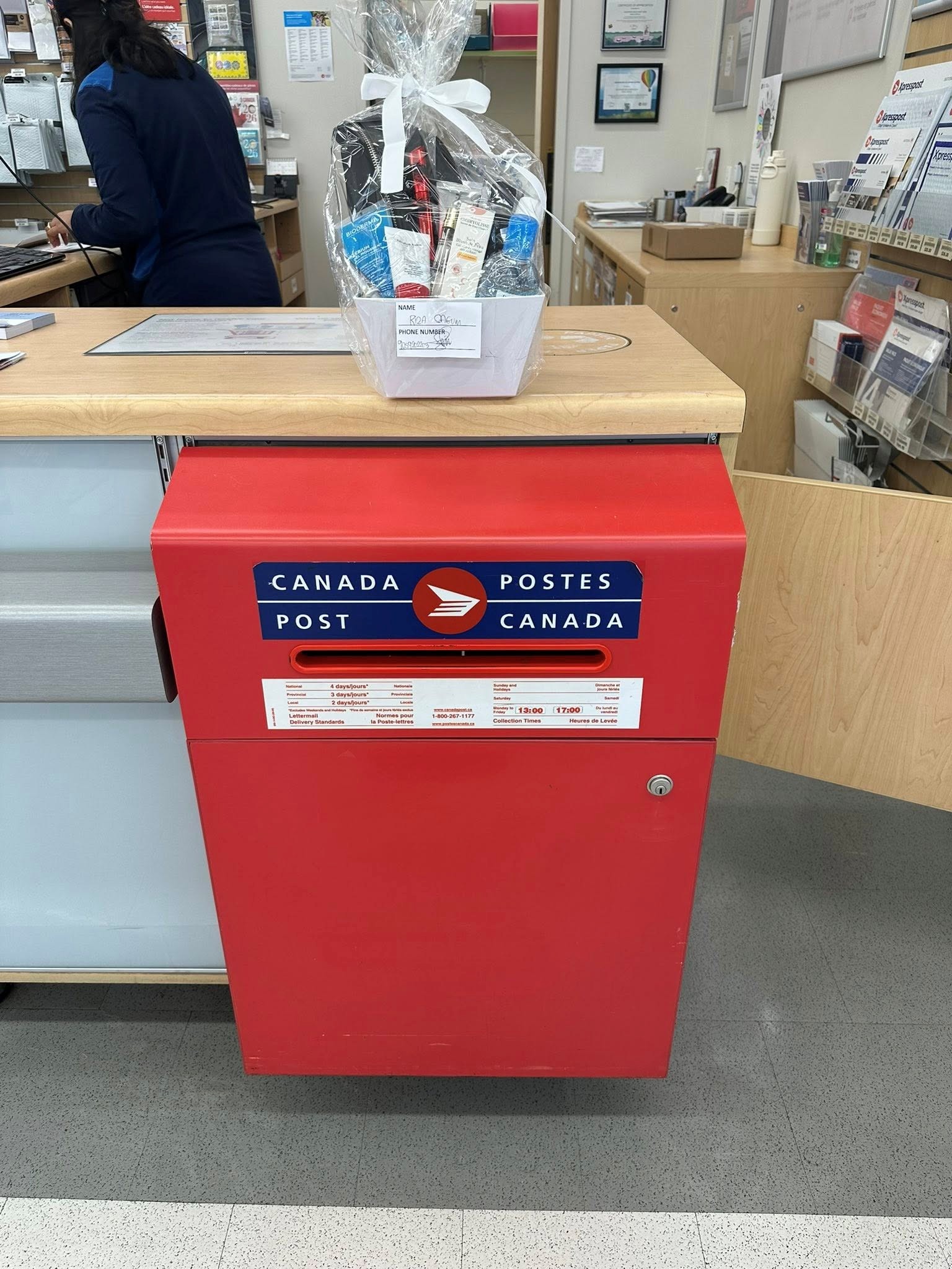 9 - The Canada Times Red Canada Post mailbox at a service counter with a gift basket wrapped in cellophane on top, containing toiletries and goodies.