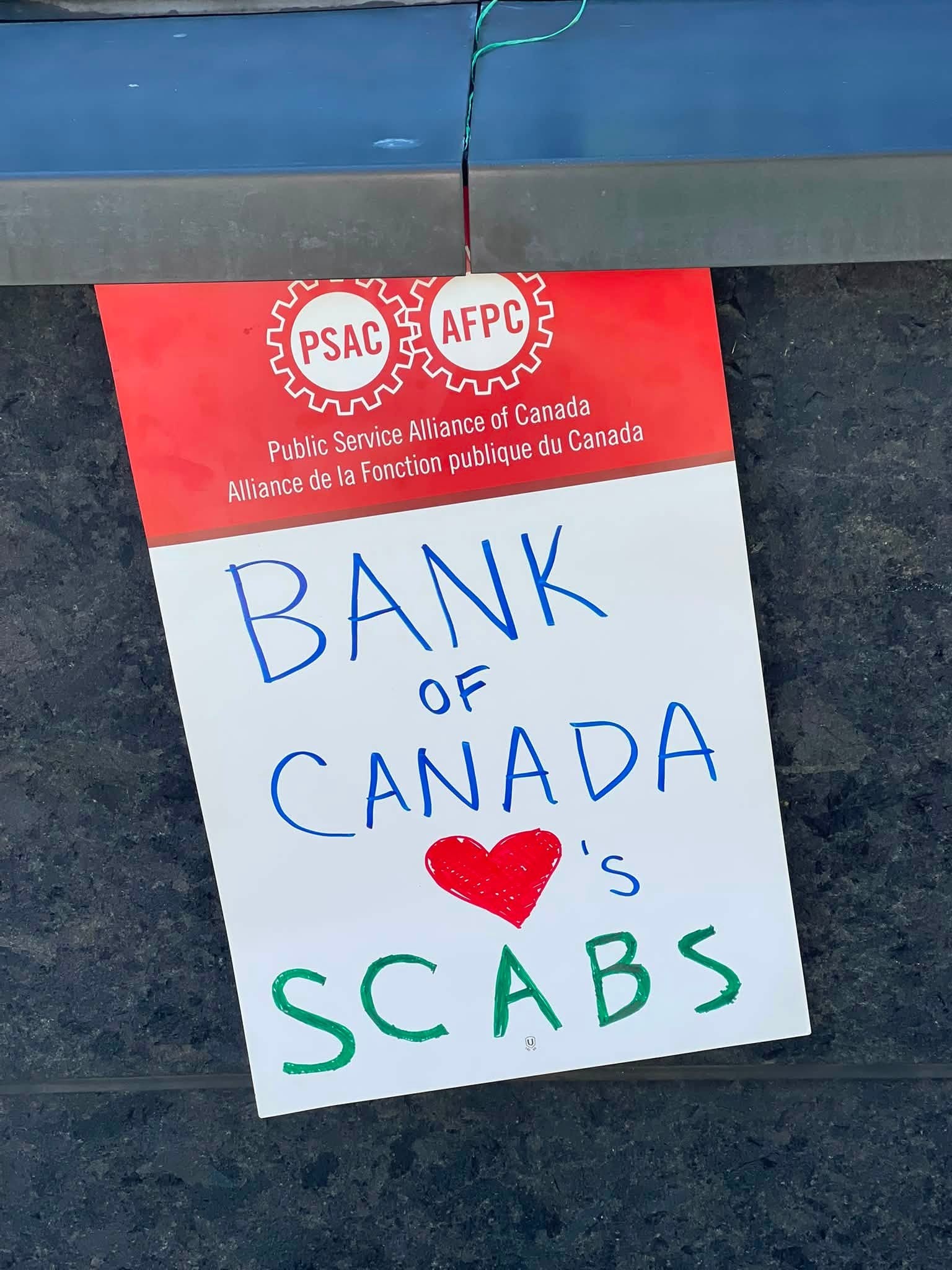 Red union poster with PSAC and AFPC logos and 'Public Service Alliance of Canada' text; below is a white sheet reading 'BANK OF CANADA' in blue with a red heart.