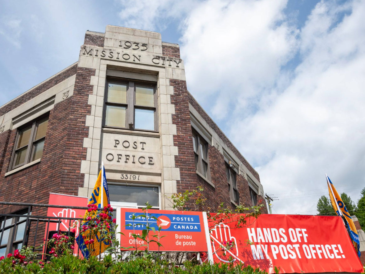 9 - The Canada Times Brick post office building with a stone-framed sign reading ‘Post Office’ and protest banners in front under a blue sky.