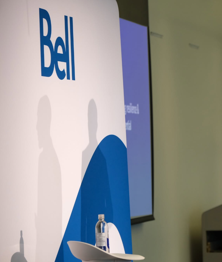 Dell event banner with large Dell logo and blue abstract shapes; two silhouette shadows on white backdrop, water bottle and plate on a podium in foreground