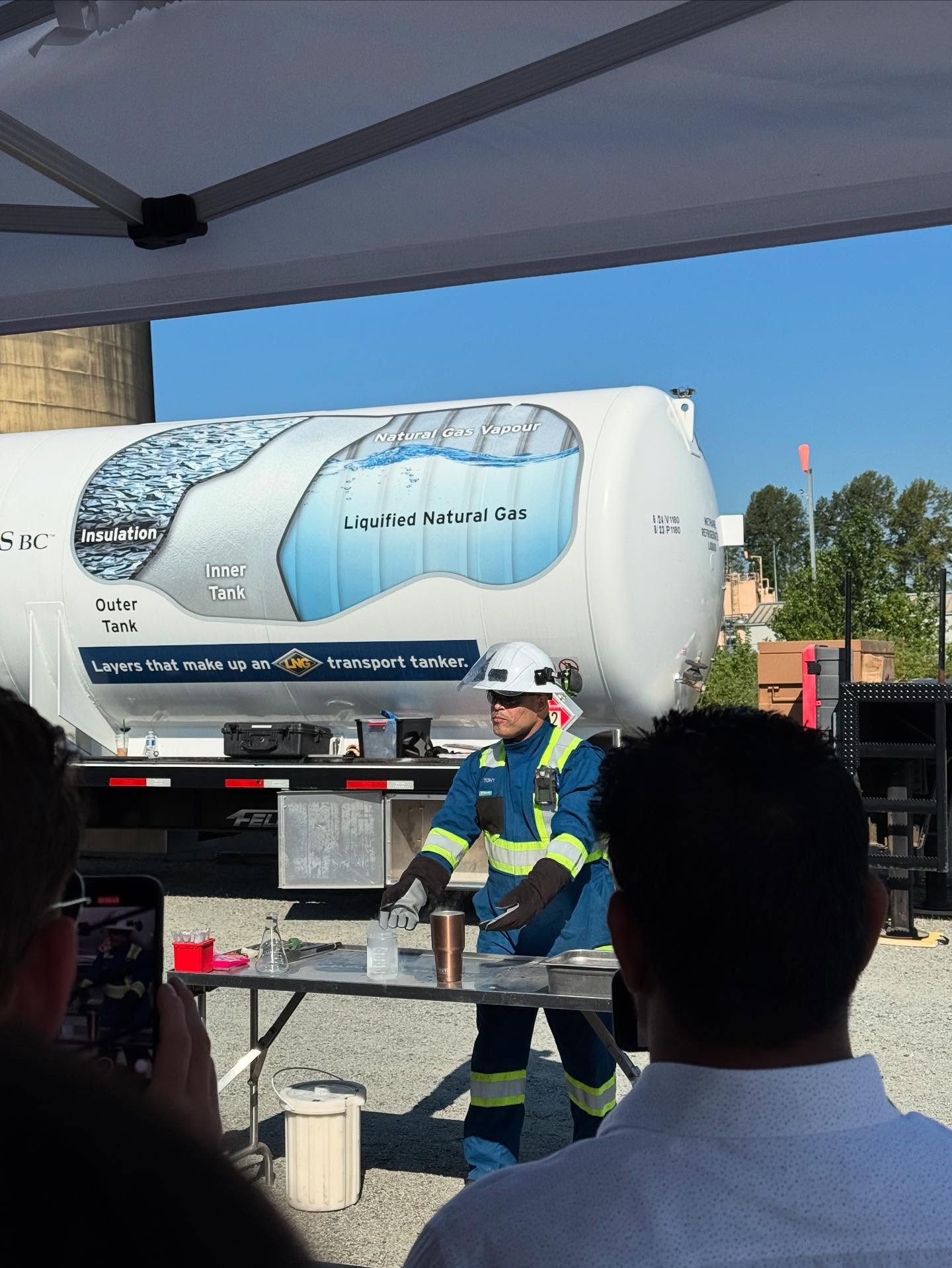 Worker in blue safety gear conducts an outdoor demonstration with lab equipment on a table, LNG tanker trailer visible in the background under a tent.