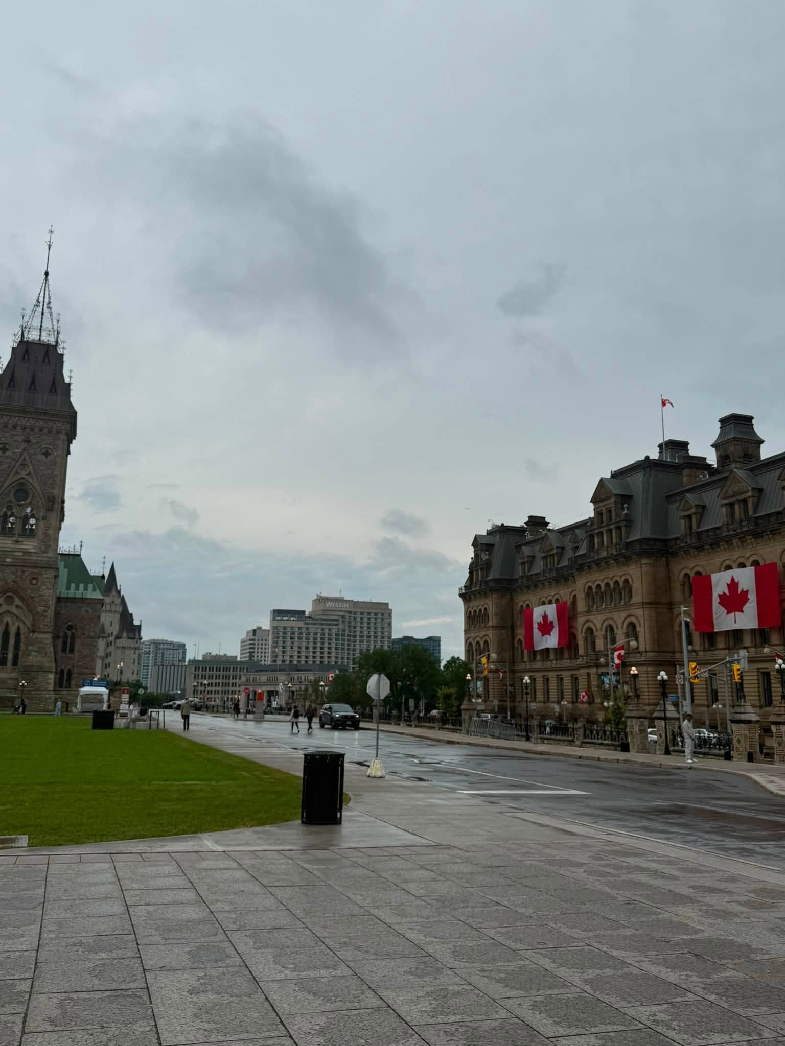 1 - The Canada Times Wide city square with historic buildings and Canadian flags along the right, a Gothic-style tower on the left, and a wet, reflective pavement under a cloudy sky.