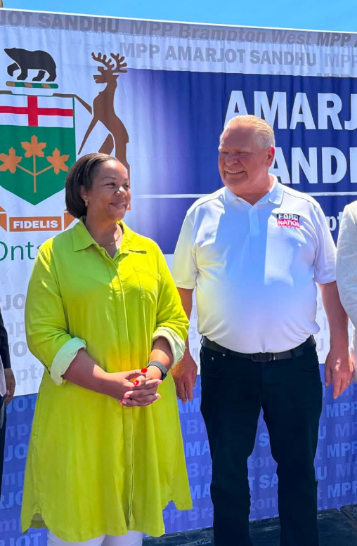 Two adults smile for a photo in front of a blue event banner; woman wears a lime-green coat, man wears a light blue polo shirt with a badge on it.