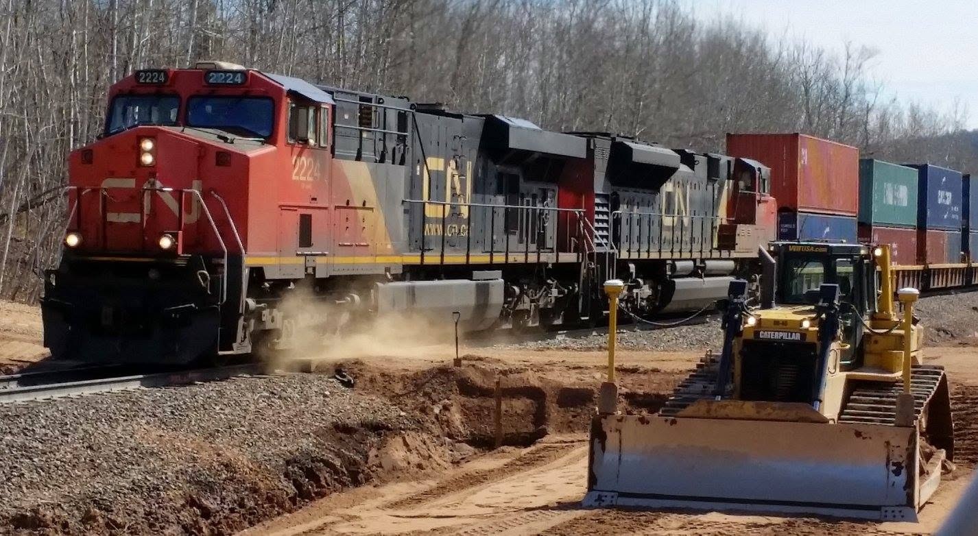 Red diesel locomotive hauling freight cars on a new track, with a bulldozer grading the adjacent earth and dust near the rails.