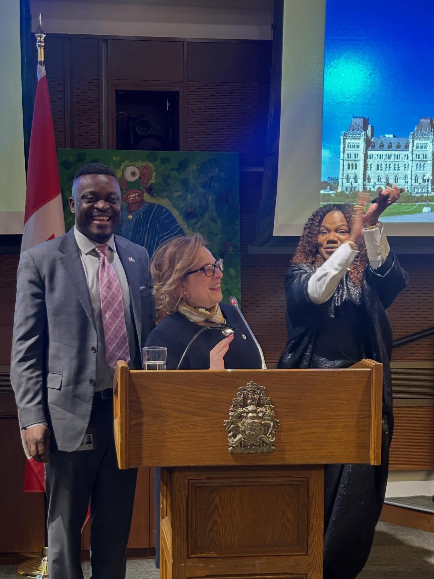 11 - The Canada Times Three people at a wooden podium during a formal event; man in gray suit and pink tie stands beside a woman with glasses, while another woman raises her hands in celebration, with a Canadian flag and a mural in the background.