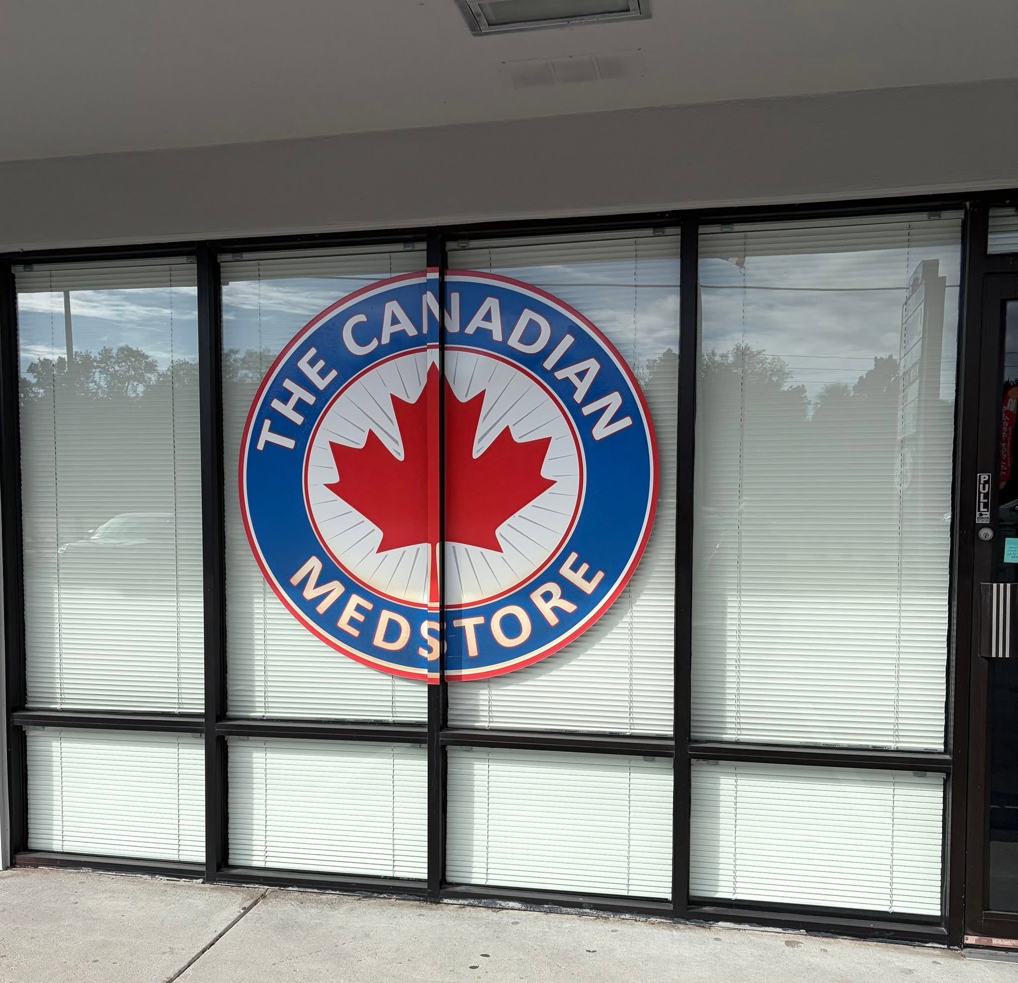 Storefront with a large circular Canada-themed logo on the glass doors, blinds in the windows, and a concrete sidewalk in front.