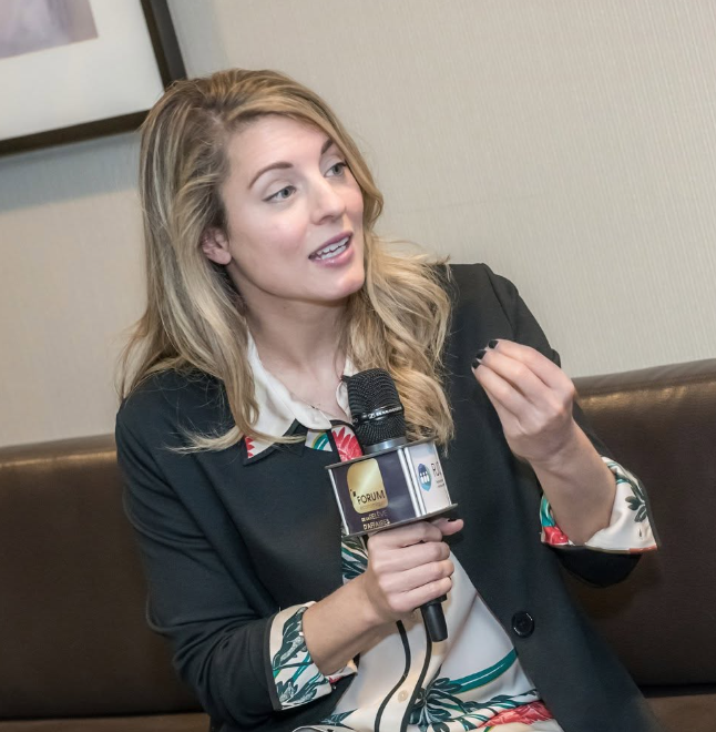 11 - The Canada Times Blonde woman in a dark blazer sits on a brown couch, holding a microphone and speaking in an interview setting.