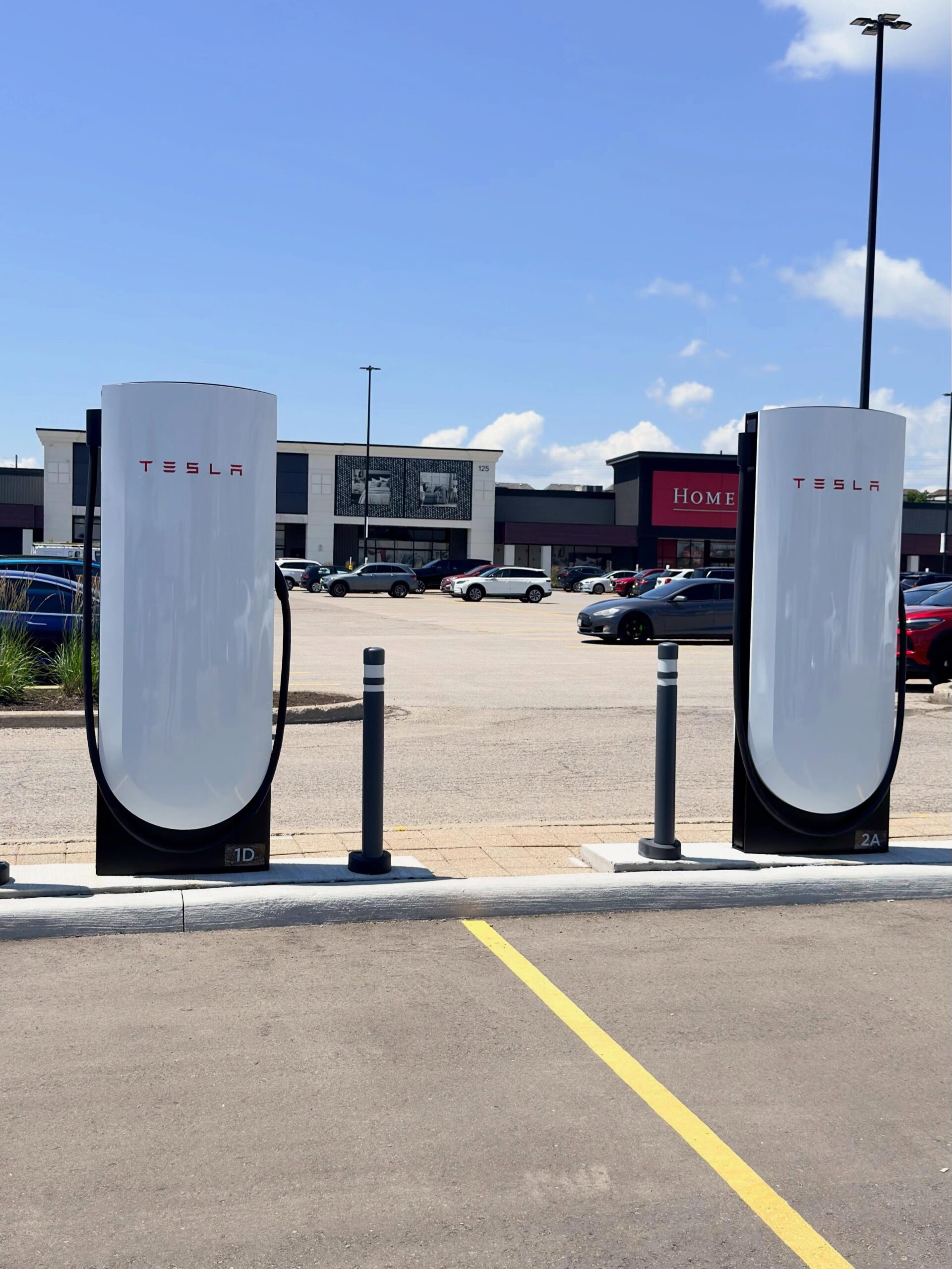 Two Tesla electric vehicle charging stations in a parking lot, with cables hanging from white units under a clear blue sky