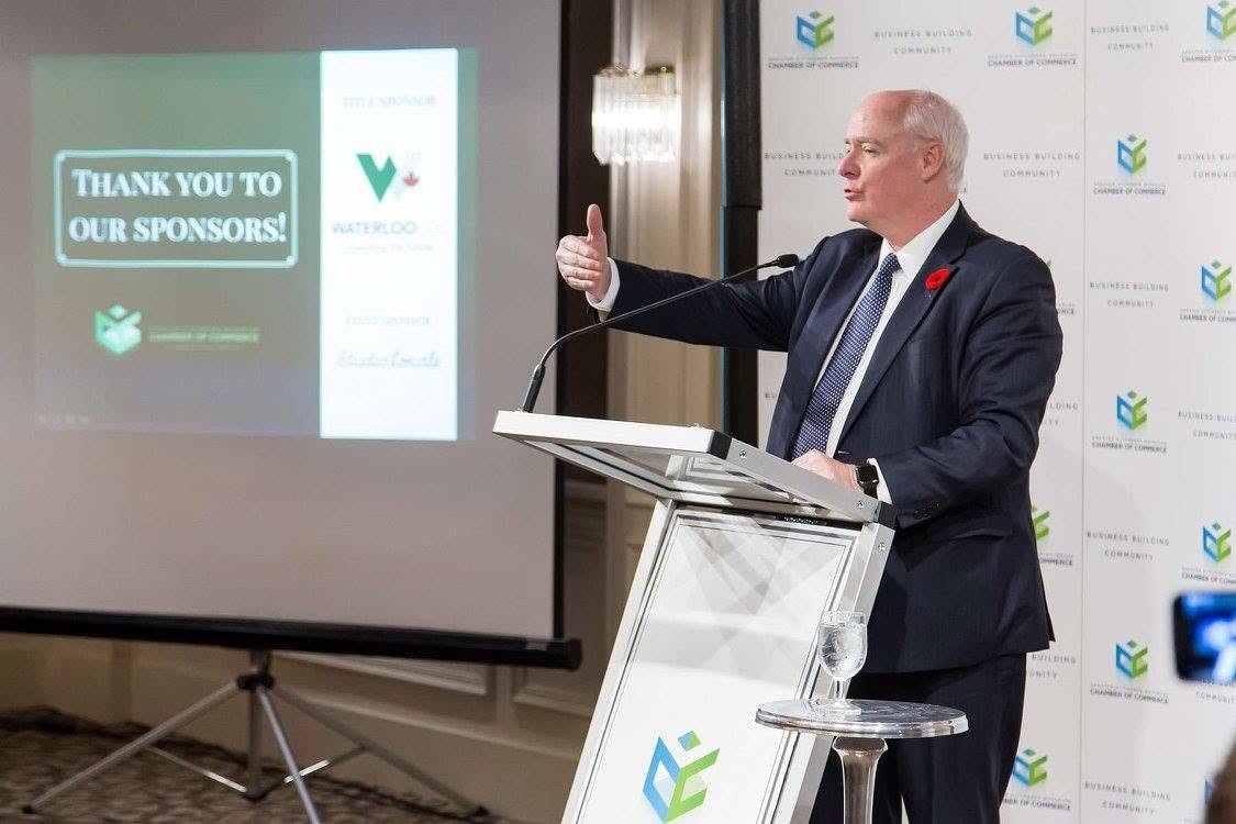 12 - The Canada Times Male speaker in a suit at a podium giving a thumbs-up, with a screen thanking sponsors in the background.