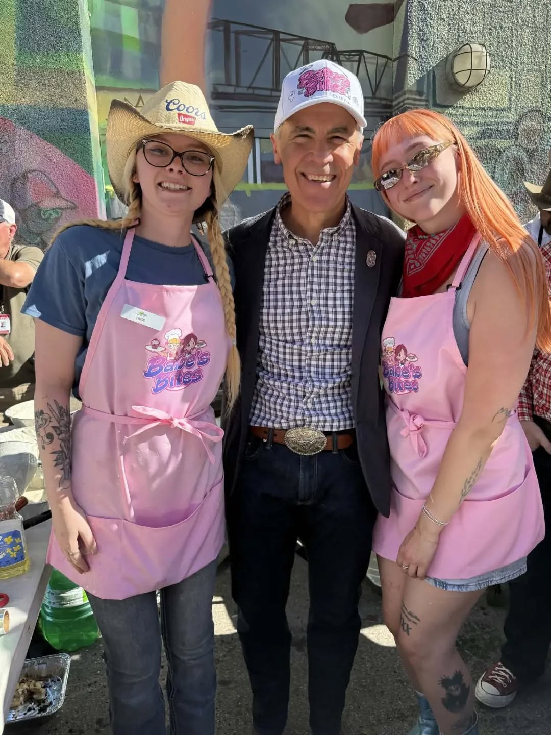 13 - The Canada Times Three people pose together outdoors in front of a colorful mural; two women wear pink aprons and name tags, with a man in a checkered shirt and blazer between them, smiling.