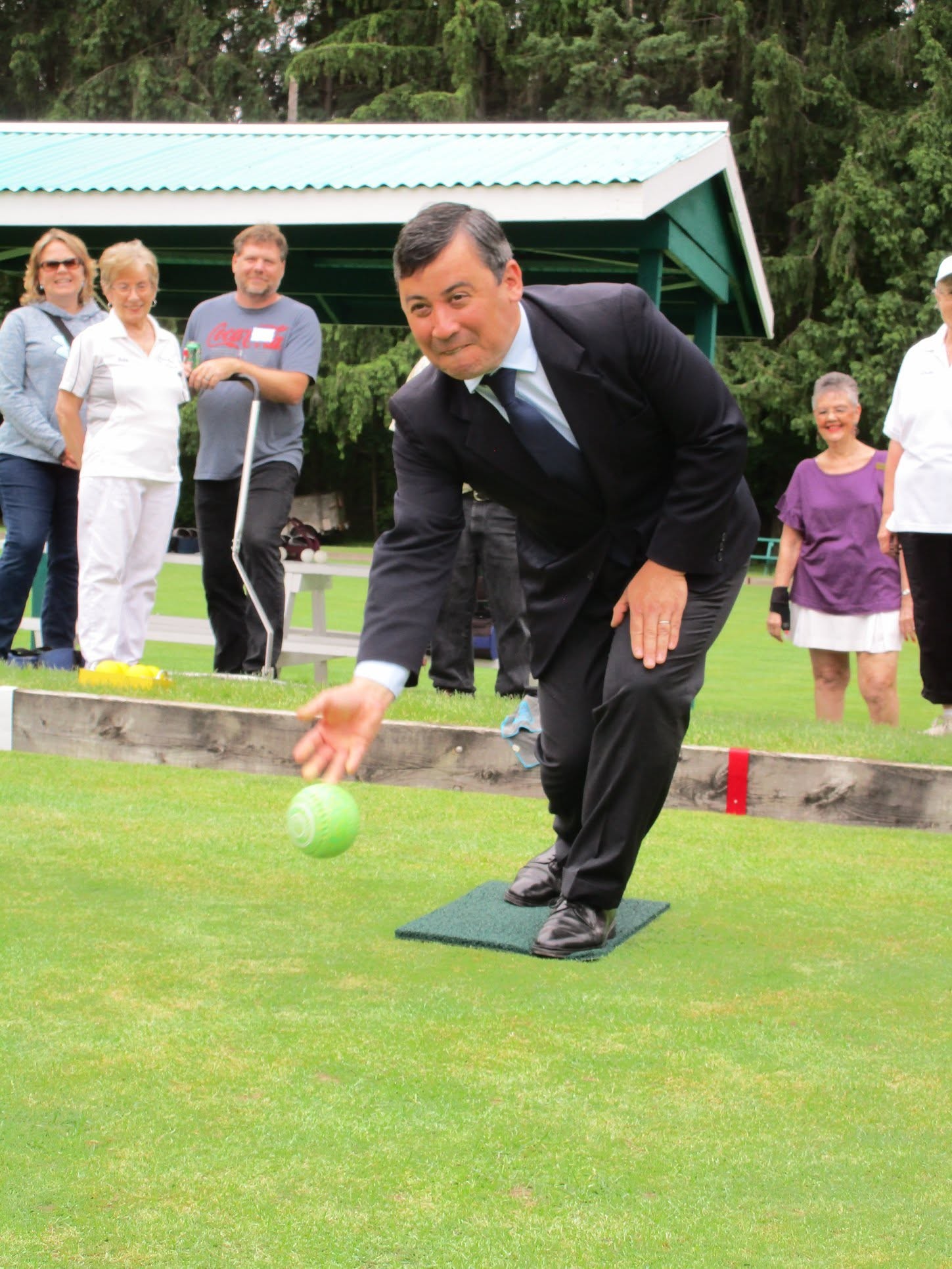 13 - The Canada Times Man in a dark suit bending to roll a lime-green ball on a grassy lawn, with several observers standing behind a wooden barrier.