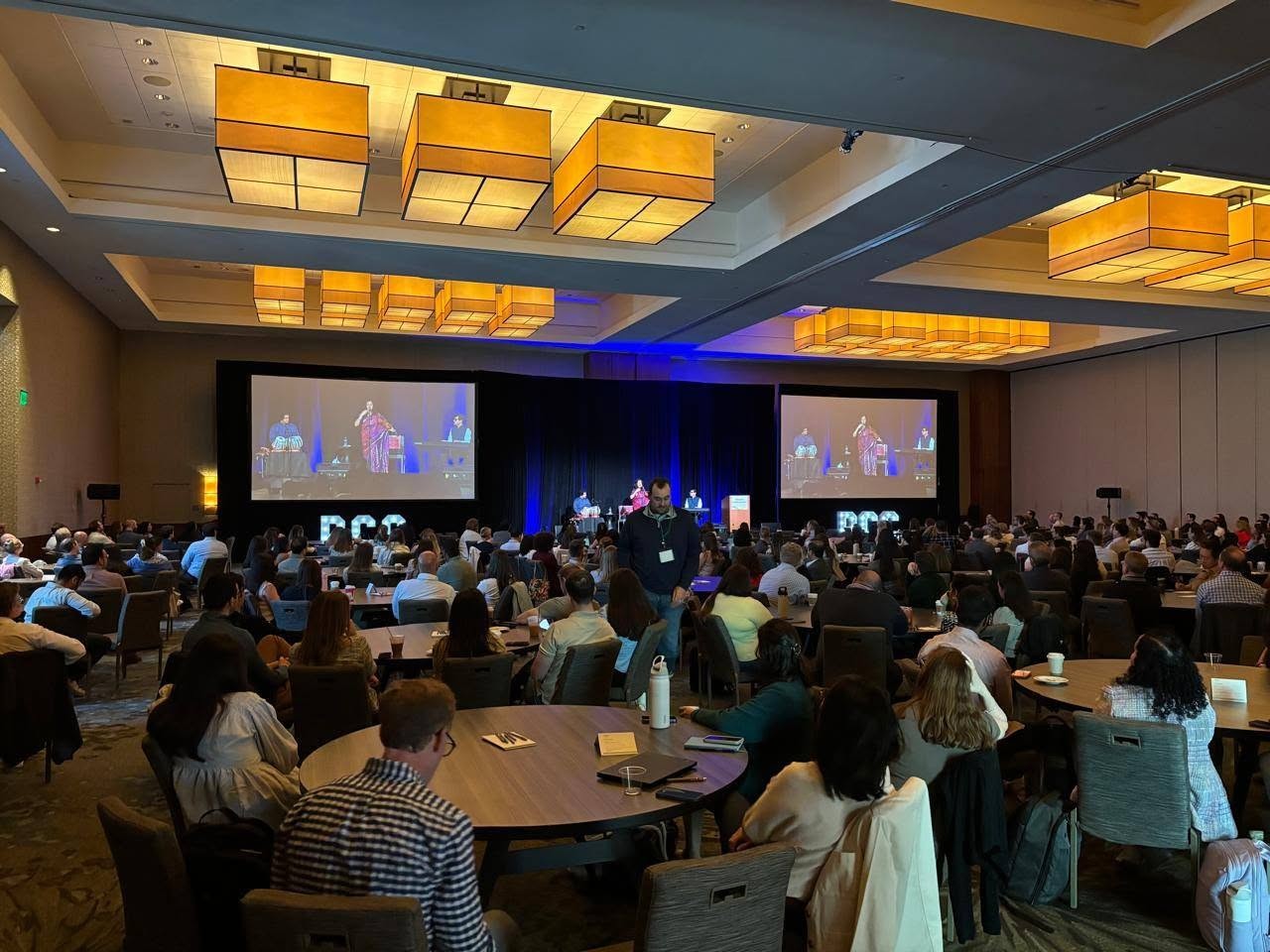 2 - The Canada Times Large conference hall with a stage and two big screens showing speakers; audience seated at round tables watching a presentation on stage.
