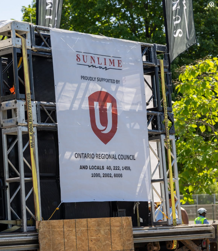2 - The Canada Times Event banner on a metal scaffold reading 'Ontario Regional Council' with a red shield logo, plus smaller text about locals, outdoors with trees in the background and a worker in safety gear nearby.