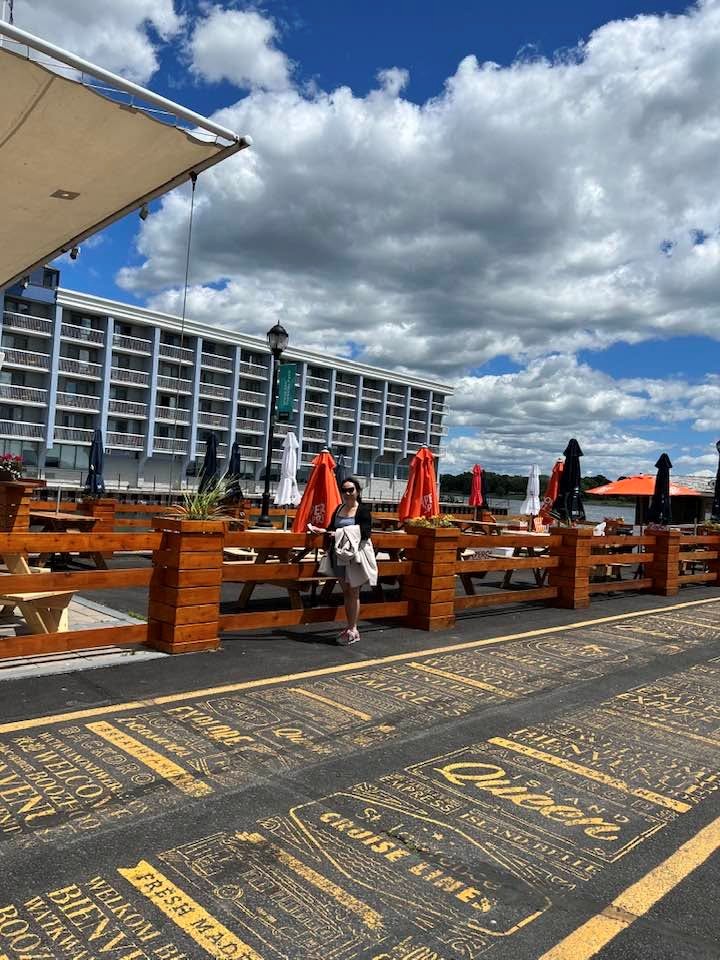 3 - The Canada Times Woman in a light jacket stands by a wooden railing on a waterfront boardwalk, with orange umbrellas and a large hotel behind under a blue, partly cloudy sky.
