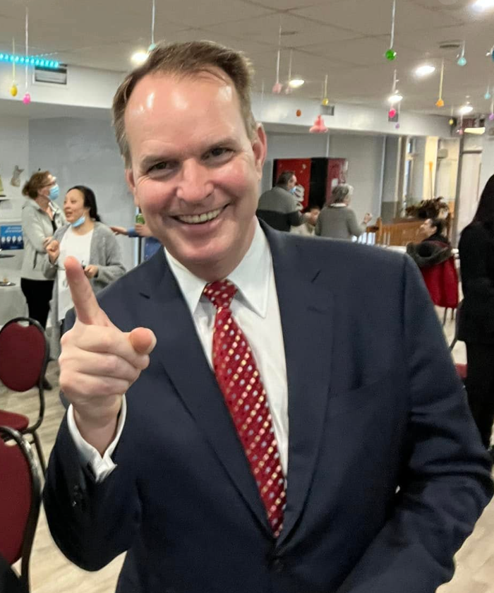 Smiling man in a navy suit and red tie points at the camera at a social event indoors.