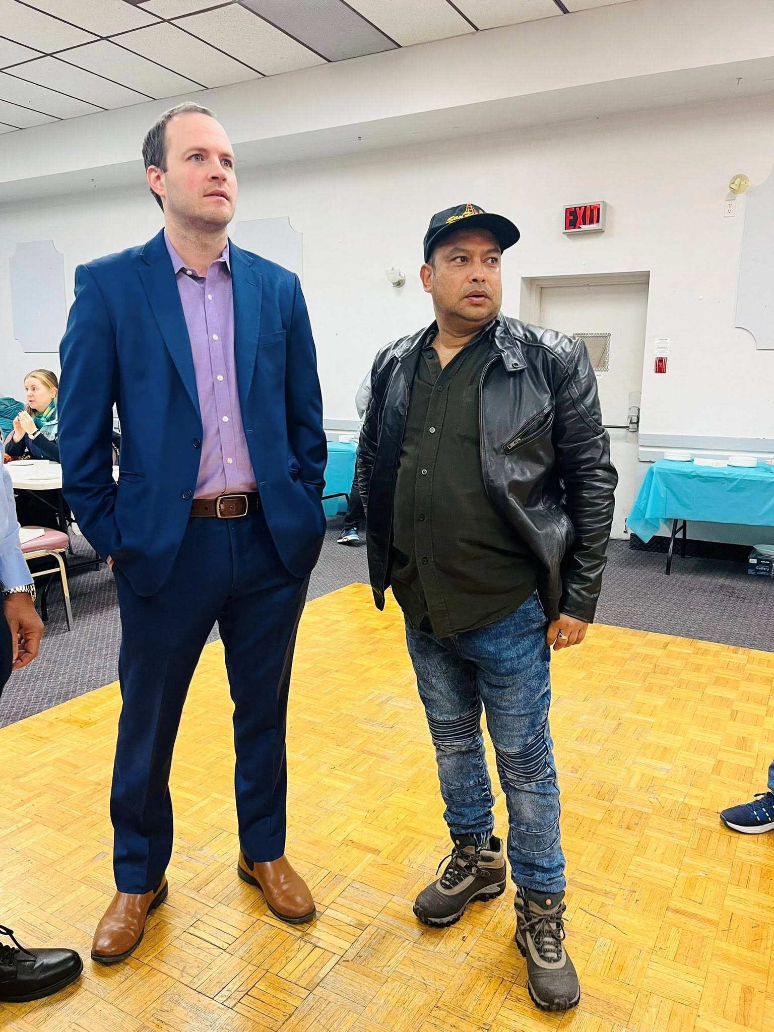 5 - The Canada Times Two men stand indoors at an event; one in a blue suit with brown shoes, the other in a black leather jacket, cap, and rugged boots, looking to the left.