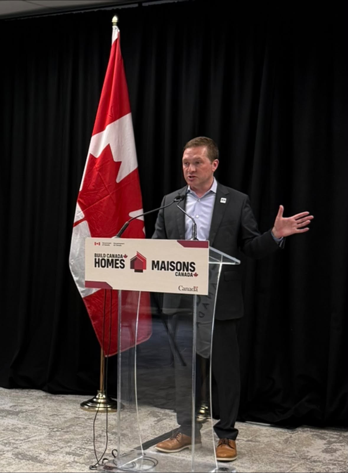 5 - The Canada Times Man in a dark suit giving a speech at a clear podium, with a large Canadian flag behind him and event signage reading 'Build Canada Homes/Maisons Canada'
