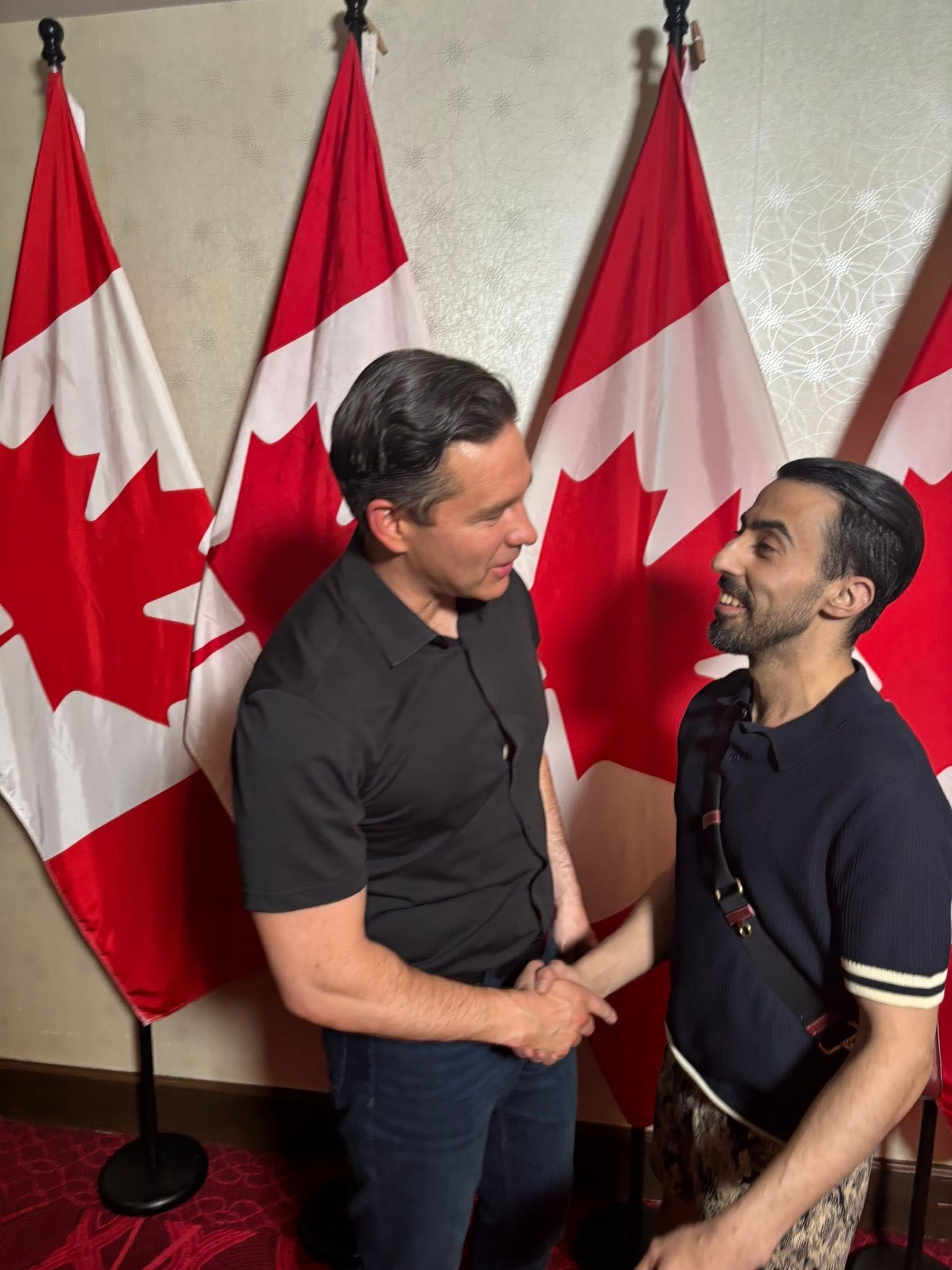 Two men shake hands and smile in front of Canadian flags at an indoor event.